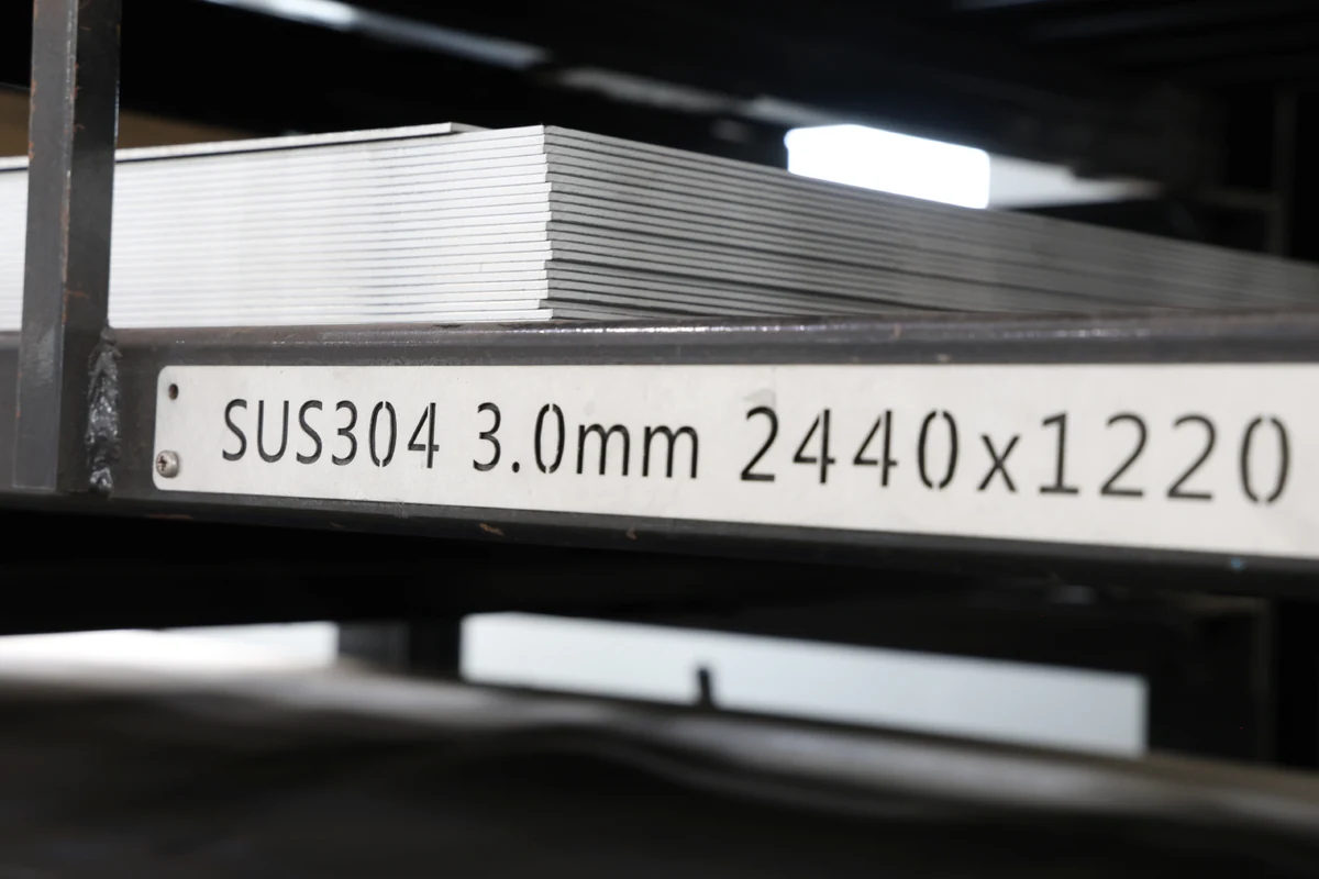 Rack label reading SUS304 3.0mm 2440x1220 below a stack of stainless steel sheets